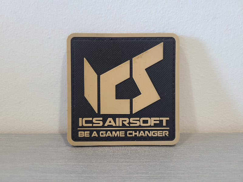 Square black and tan PVC morale patch with large ICS logo and Be a Game Changer text by DISTRICT AIRSOFT ICS airsoft PVC patch