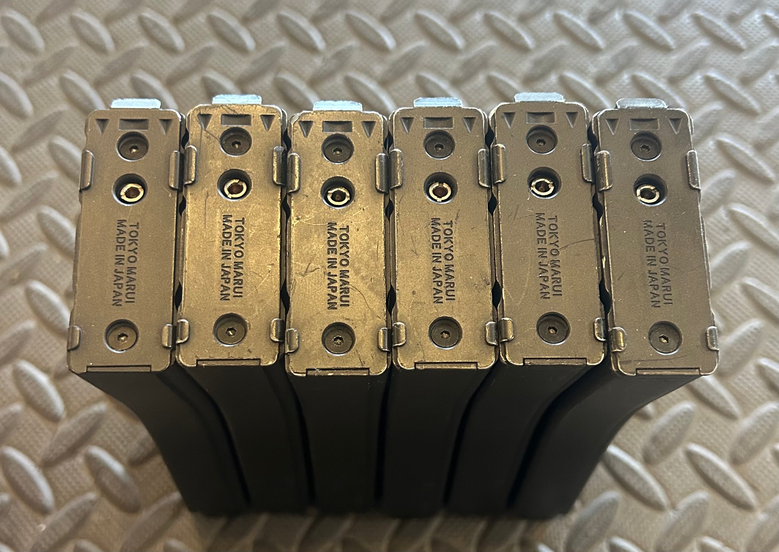 Six Tokyo Marui M4 MWS GBBR magazines stacked on a textured surface for airsoft enthusiasts